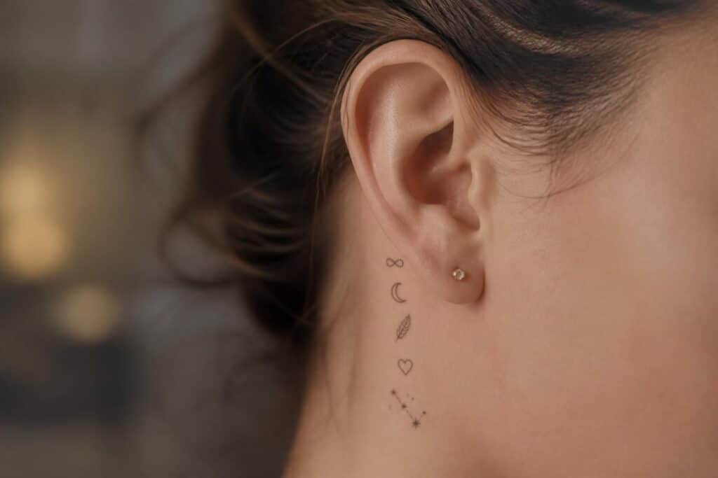 Petits tatouages alignés derrière une oreille : infini, croissant de lune, plume, cœur et étoiles